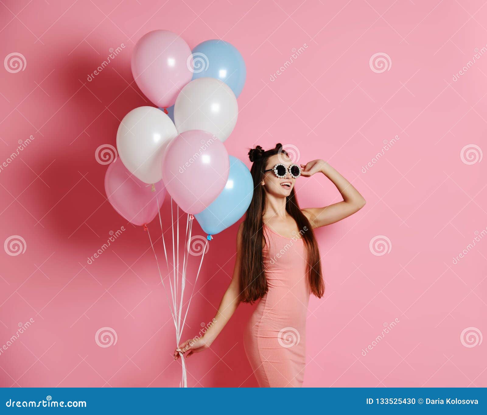 Close-up da posição loura bonito da menina em um estúdio, sorrindo extensamente e jogando com baloons azuis e cor-de-rosa. Jovem mulher bonita de sorriso nos óculos de sol à moda que têm o divertimento com os balões de ar da cor pastel em um estúdio sobre o fundo cor-de-rosa Tenha uma festa de anos do divertimento