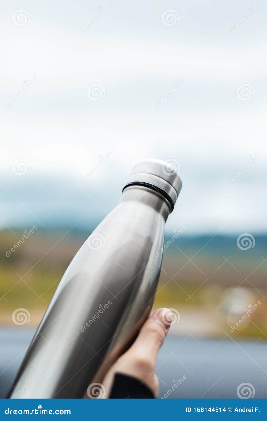 close-up of female hand holding a steel reusable thermal water bottle on background of sky.