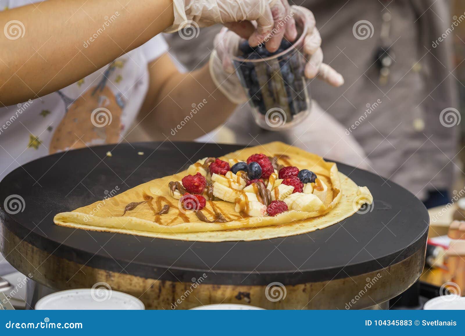 close-up of hands of cook in gloves preparing crepe, pancake on frying pan with fresh banana, blueberry, raspberry