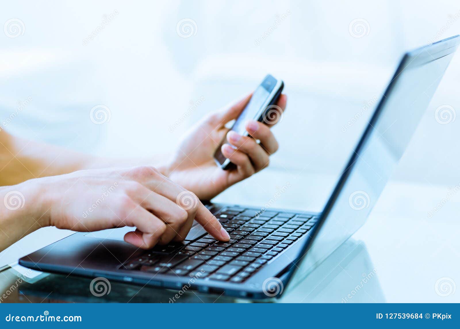 close-up of hands typing on laptop computer while using a cell phone.