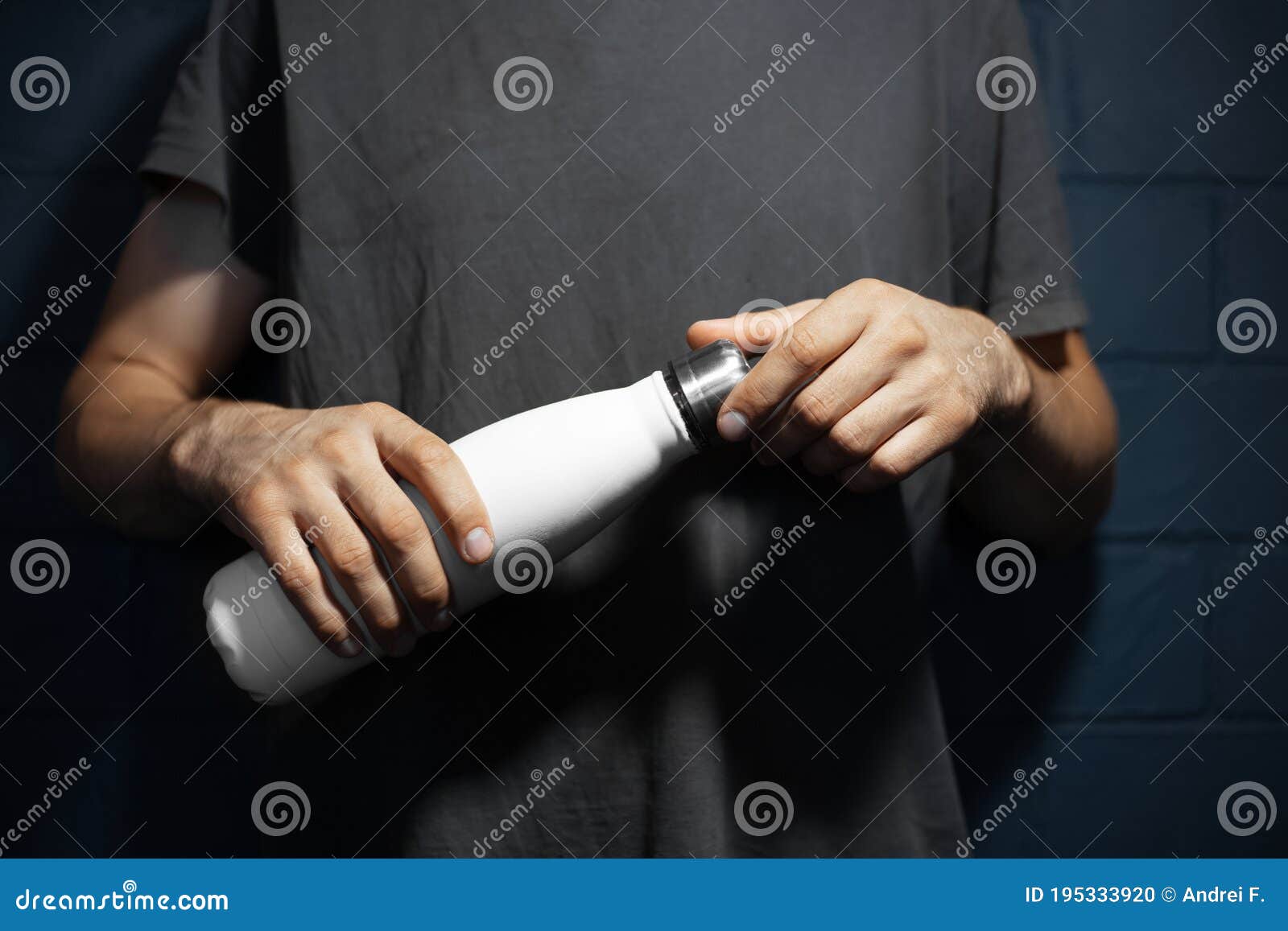close-up of male hands, openes steel thermo water bottle of white color, on the background of black brick wall.