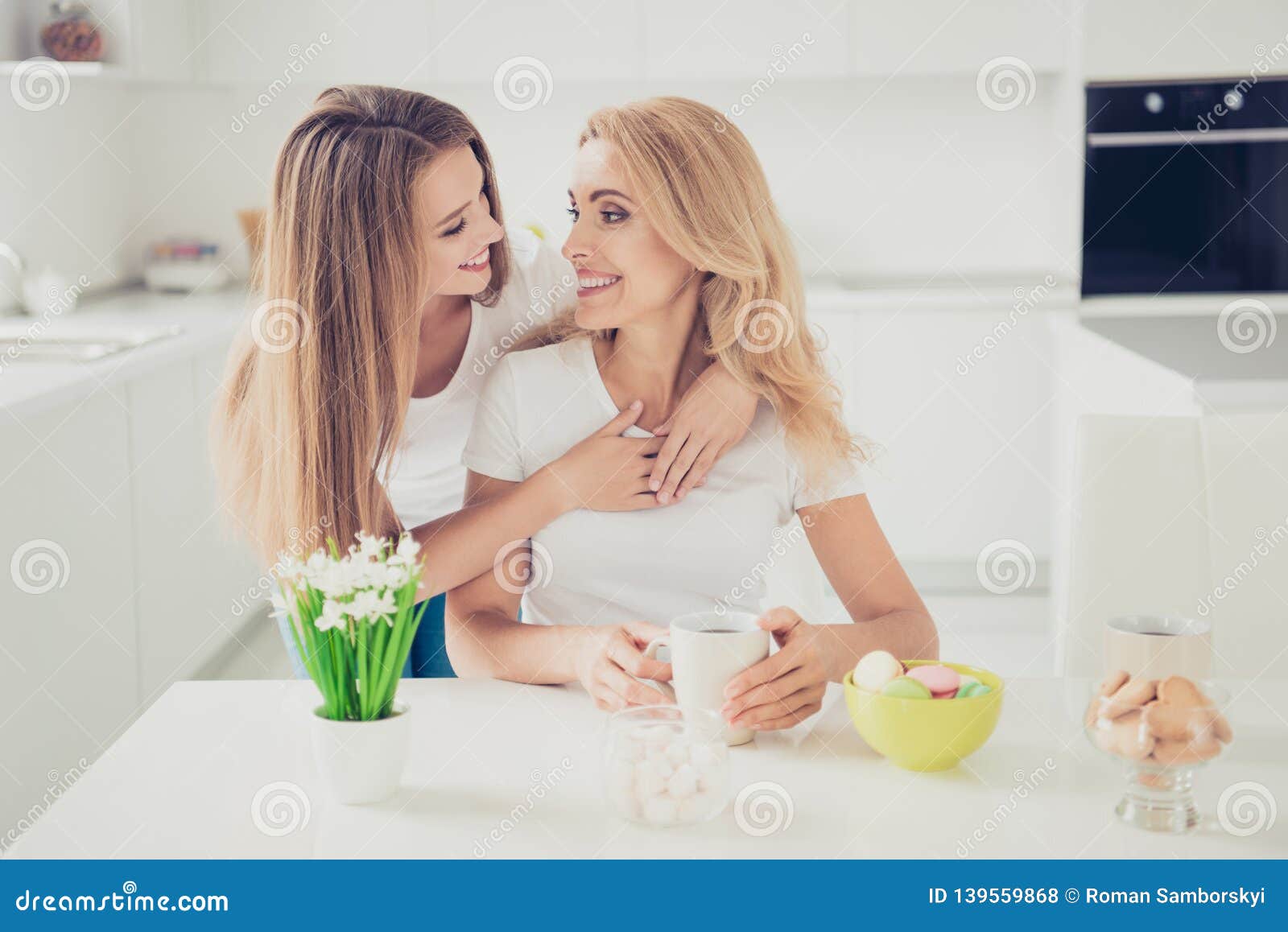 Close up photo piggy back two people mum and teenager daughter drink tea coffee family moment lovely lean on mommy look eyes women holiday wear white t-shirts jeans in bright flat kitchen.
