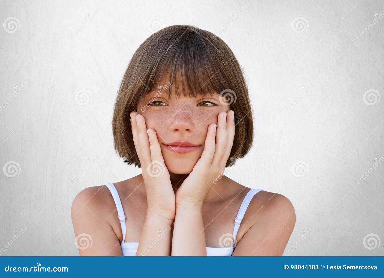 close up portrait of adorable freckled girl with bobbed hairstyle, keeping her hands on cheeks, having dreamy expression, dreaming
