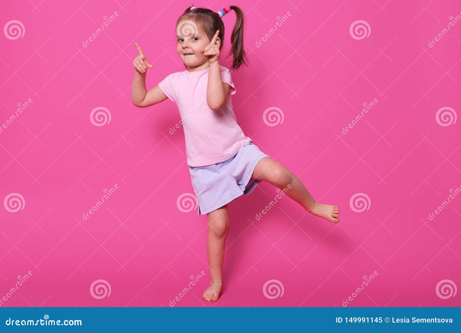 close up portrait of toddler child baby girl standing in pink casual t shirt and purple short, pointing both fore fingers up and