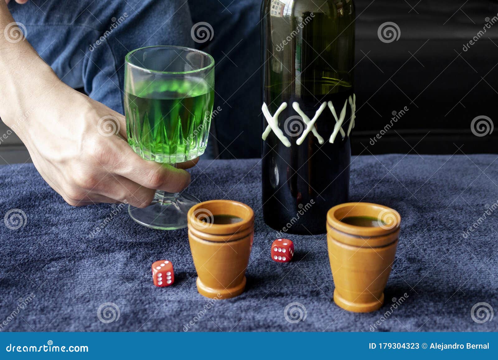 close up to a man holding a cup of green homemade liquor with two wooden cups and green artesanal alcohol bottle and dices