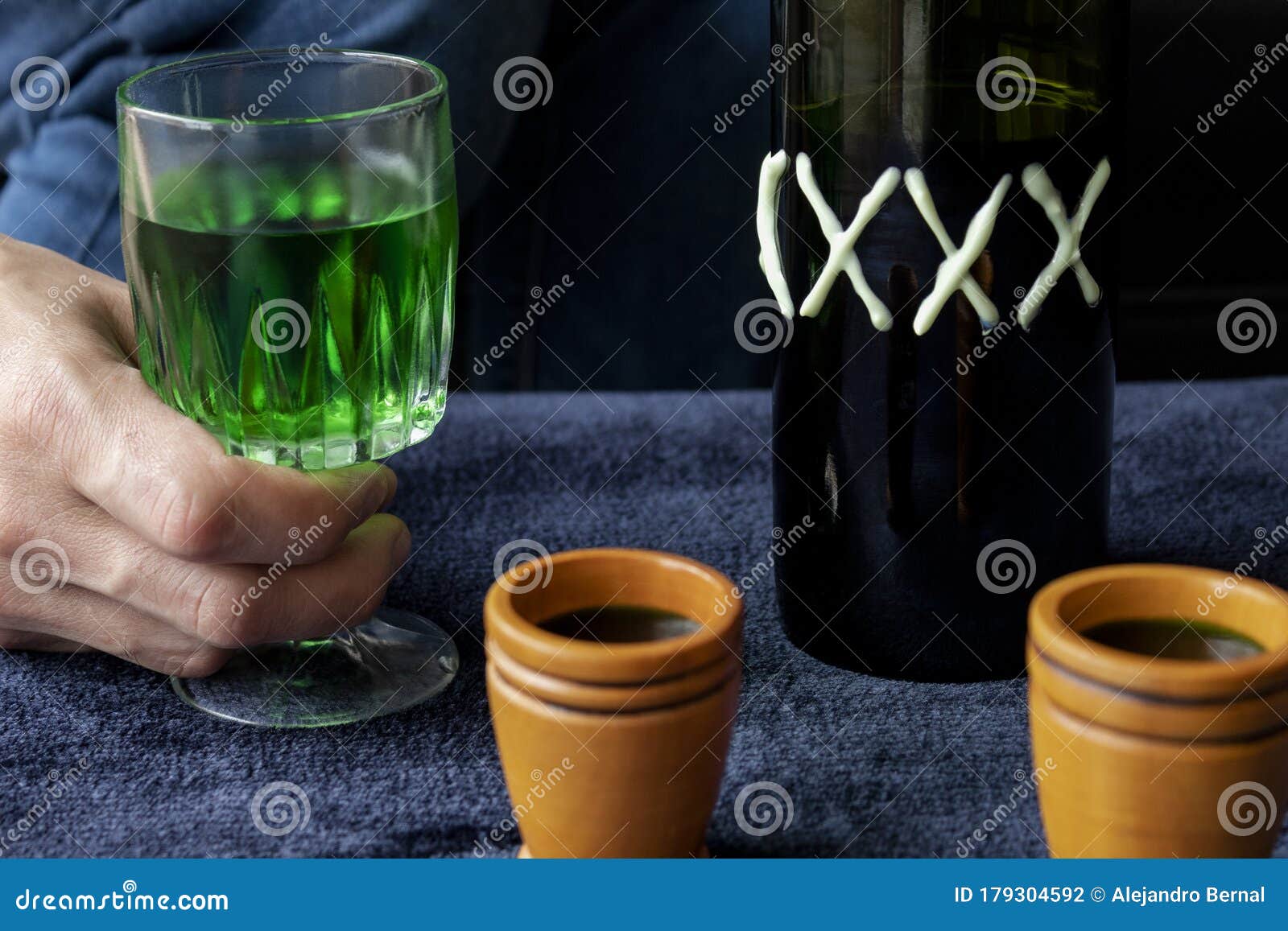 close up to a man holding a cup of green homemade liquor with two wooden cups and green artesanal bottle