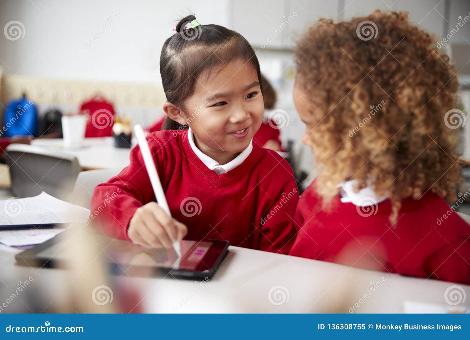 close up of two kindergarten schoolgirls wearing school uniforms, sitting at a desk in a classroom using a tablet computer and sty