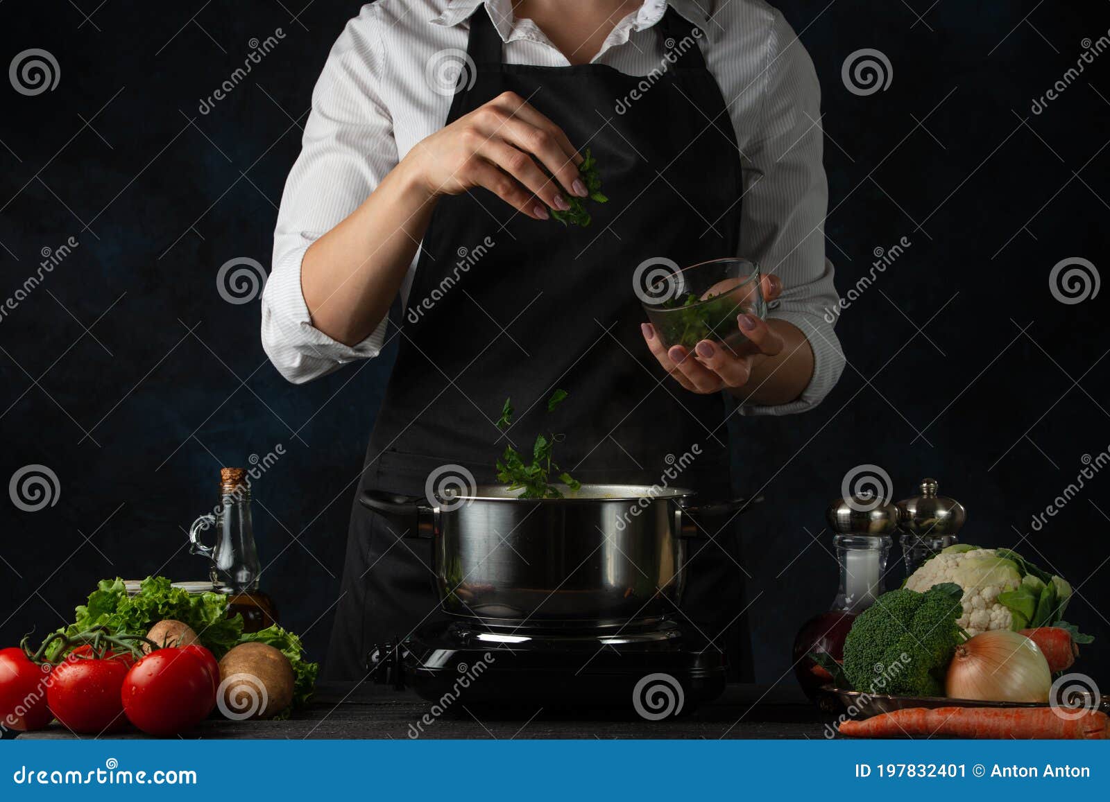 close-up view of the professional chef in black apron adding chopped parsley in boiling water for soup on dark blue background.