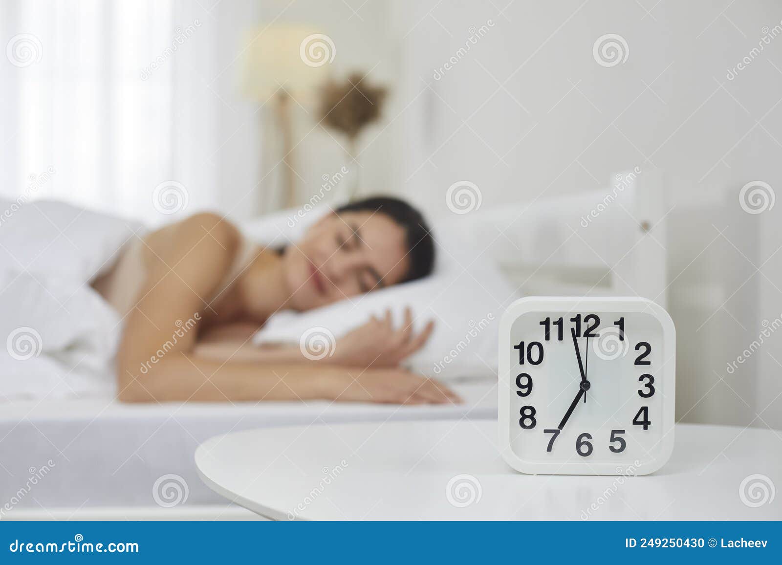 alarm clock set for 7 am on bedside table, with happy woman sleeping on bed in background