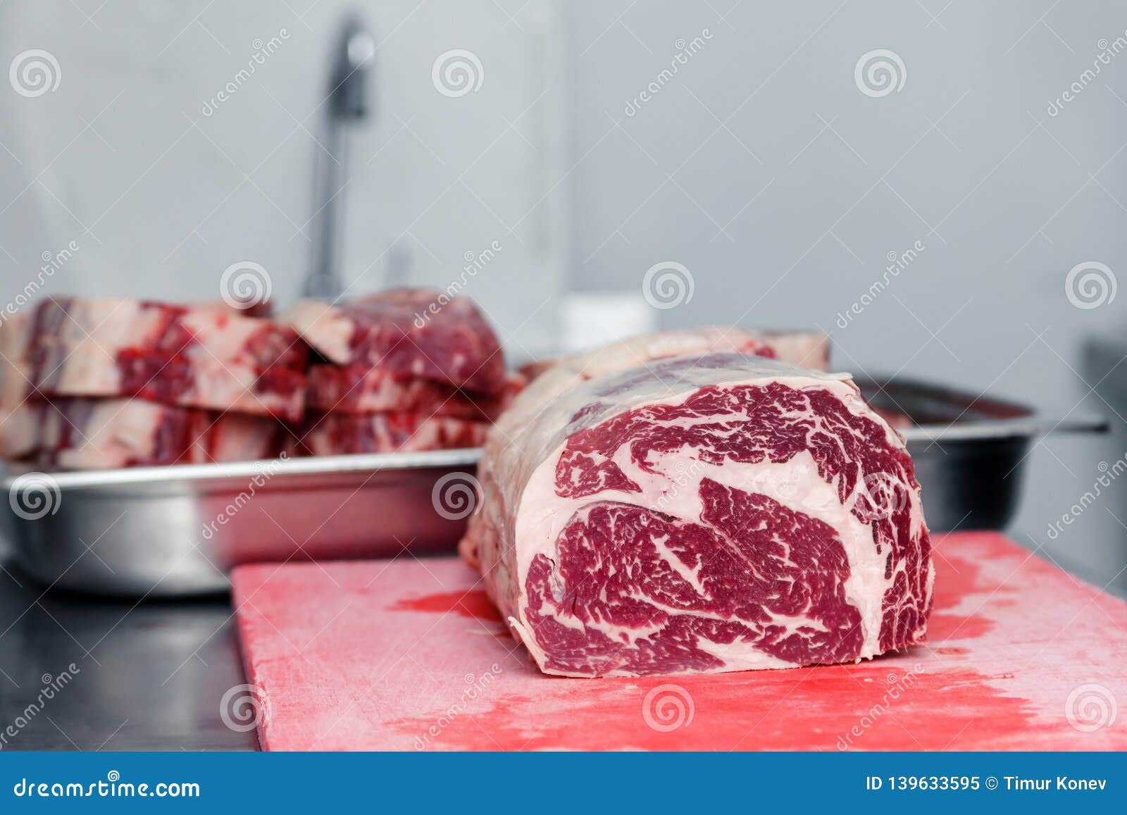 closeup cook`s hand cuts ribeye marbled beef steak with sharp knife on red plastic cutting board on metal table in restaurant