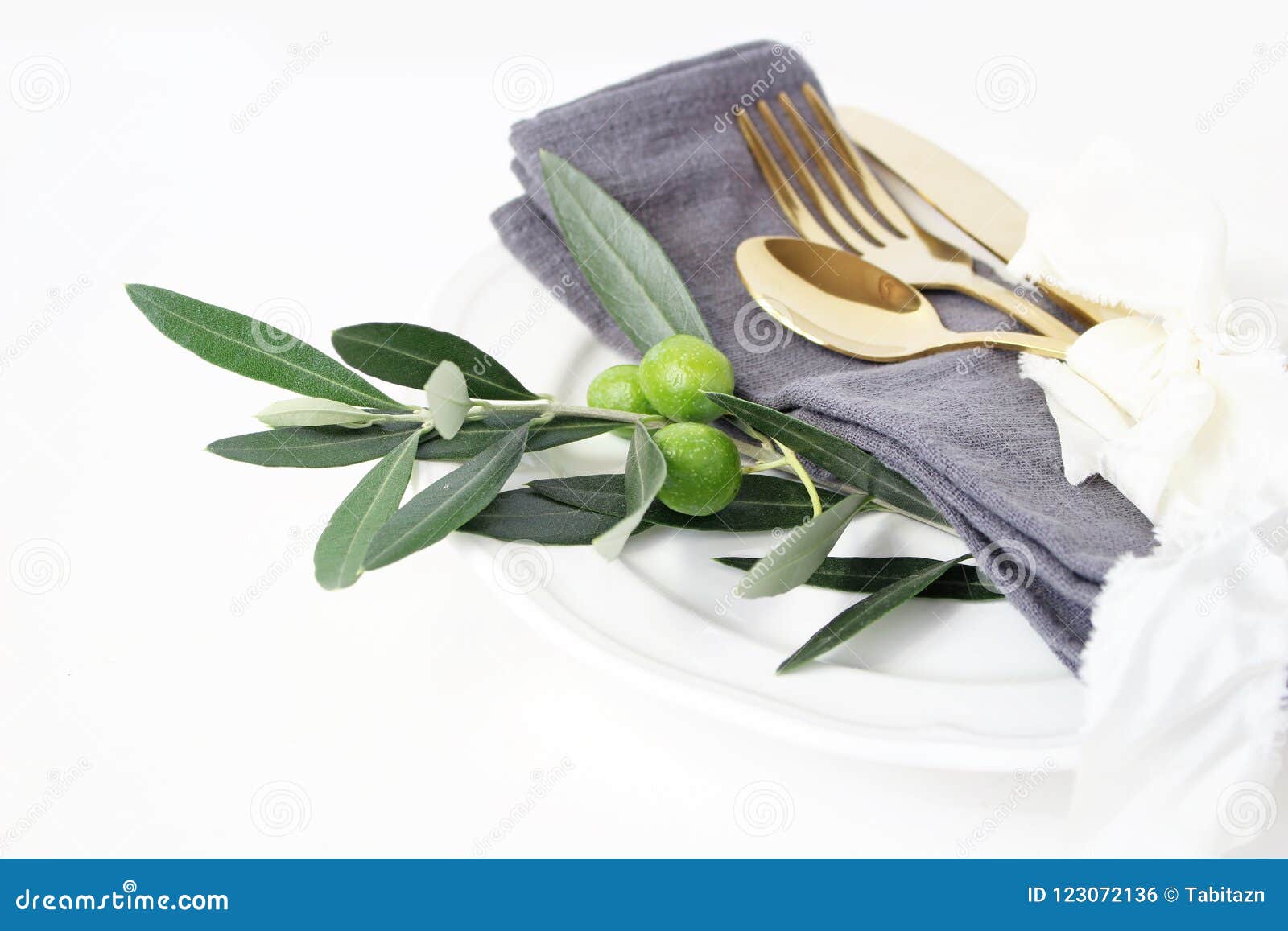 closeup of festive table summer setting with golden cutlery, olive branch, grey linen napkin, porcelain dinner plate and