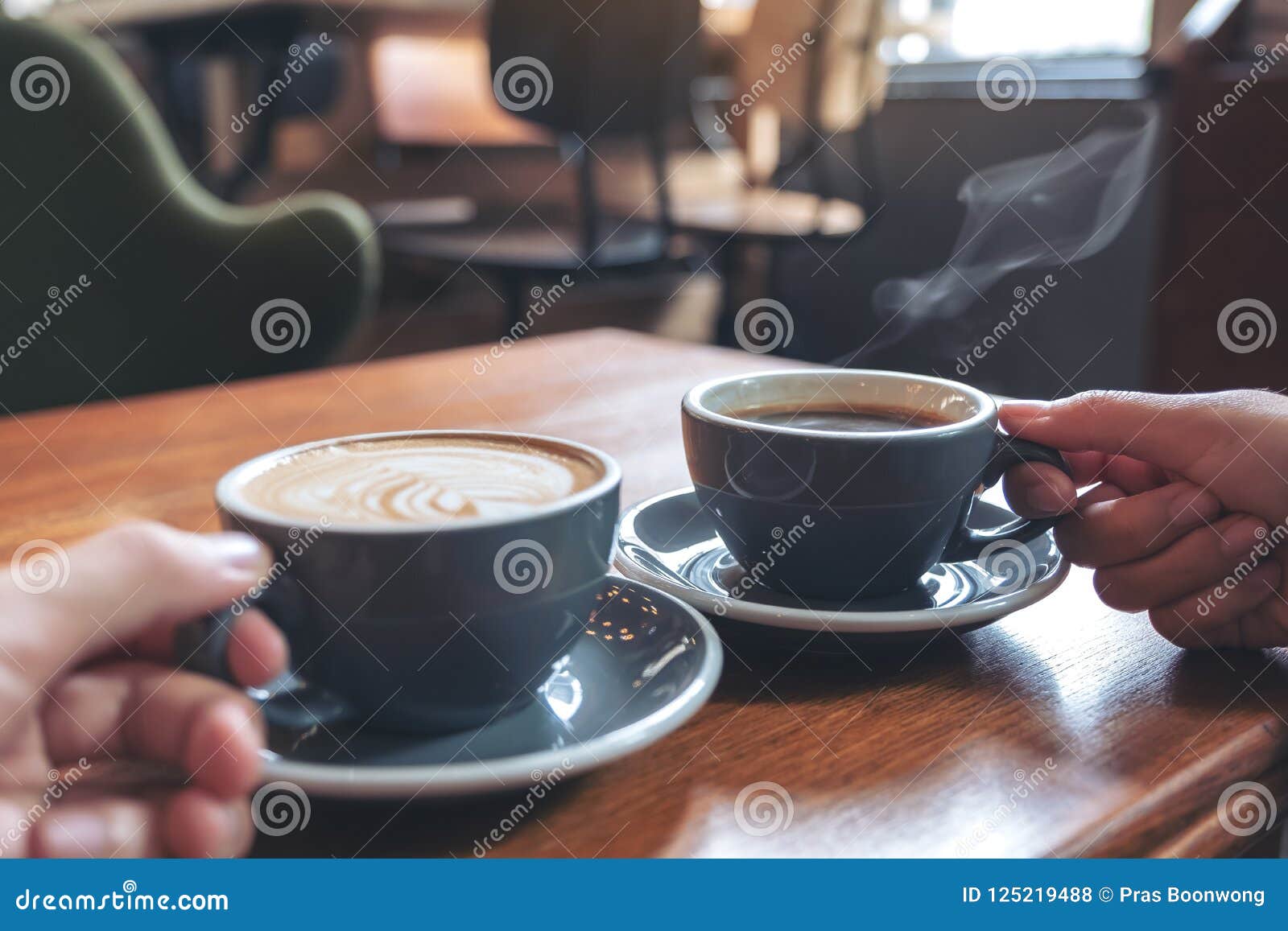 two people`s hands holding coffee and hot chocolate cups on wooden table in cafe
