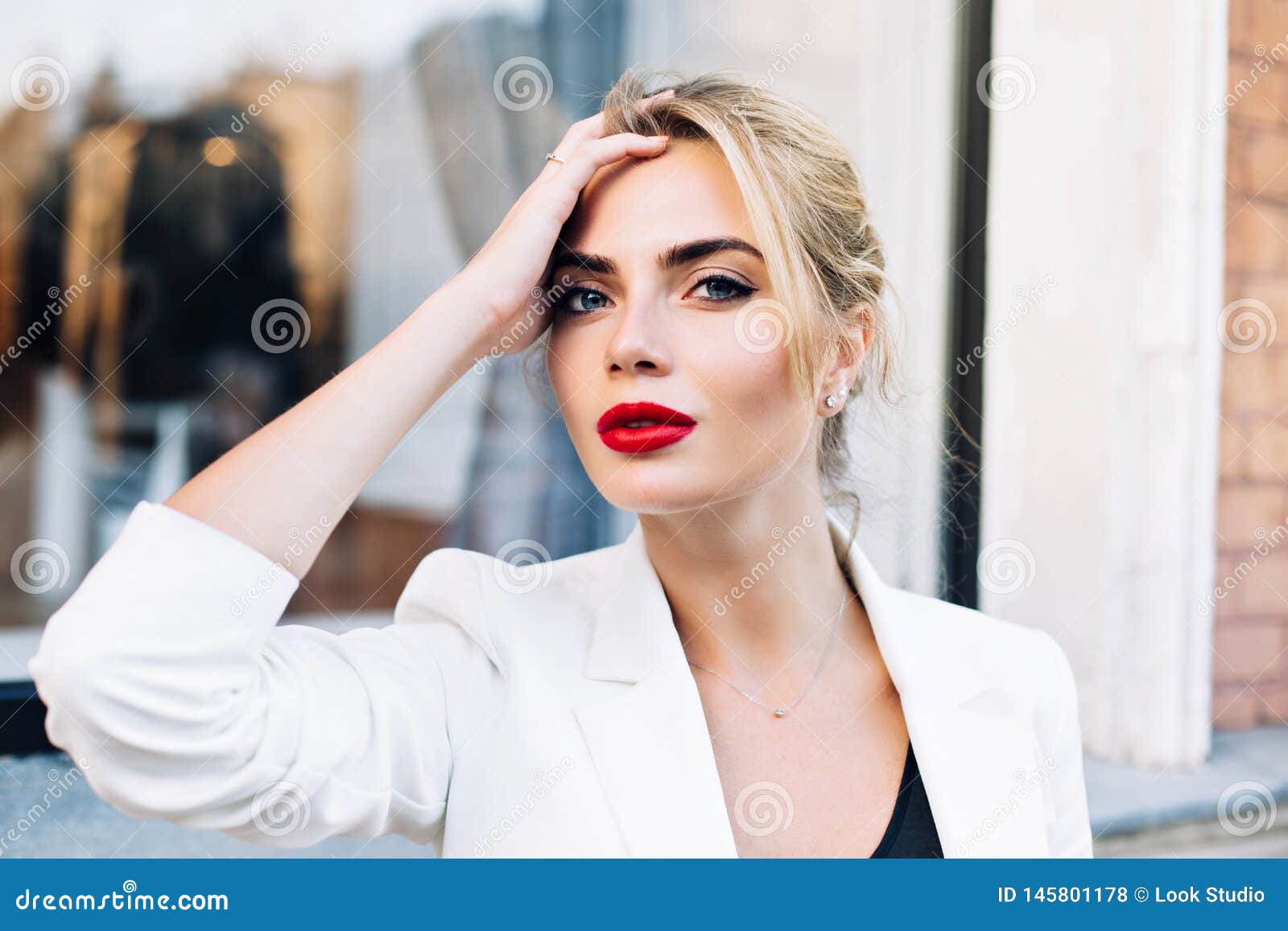 closeup portrait attractive woman with red lips on street . she wears white jacket, touching hair, looking to camera.