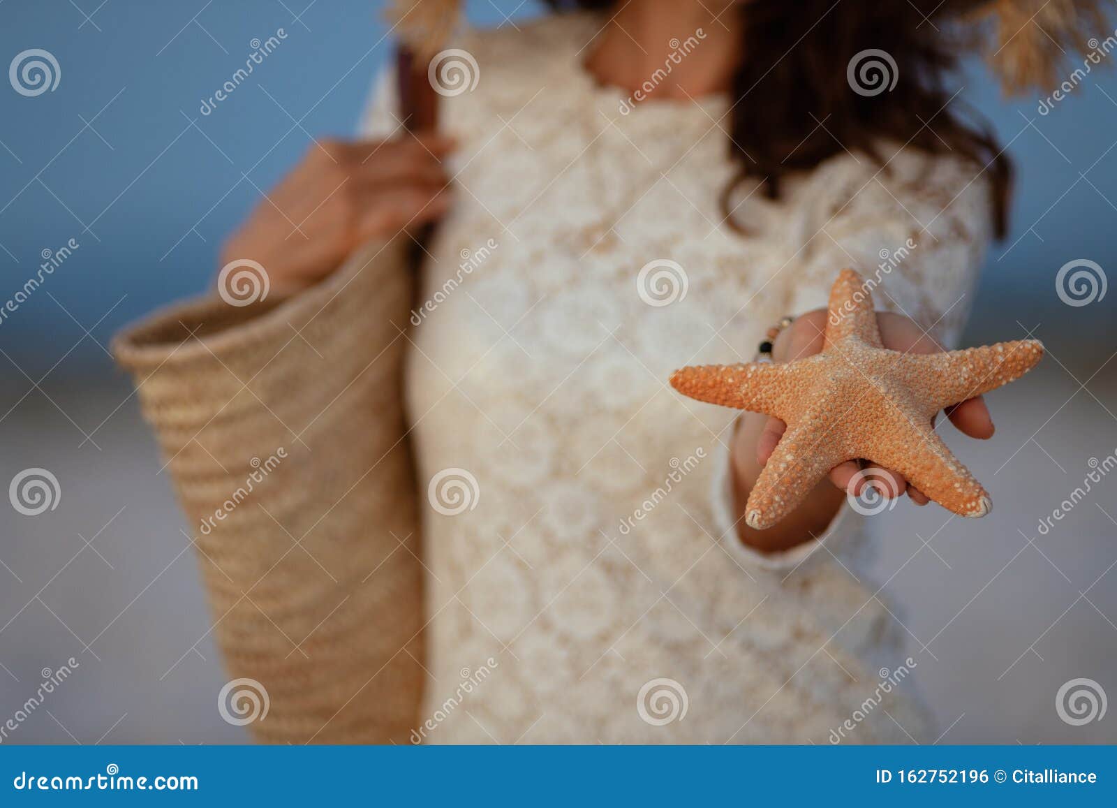 Closeup on woman on seashore at sunset showing starfish. Closeup on 40 year old woman in white dress and straw hat on the seashore at sunset showing starfish