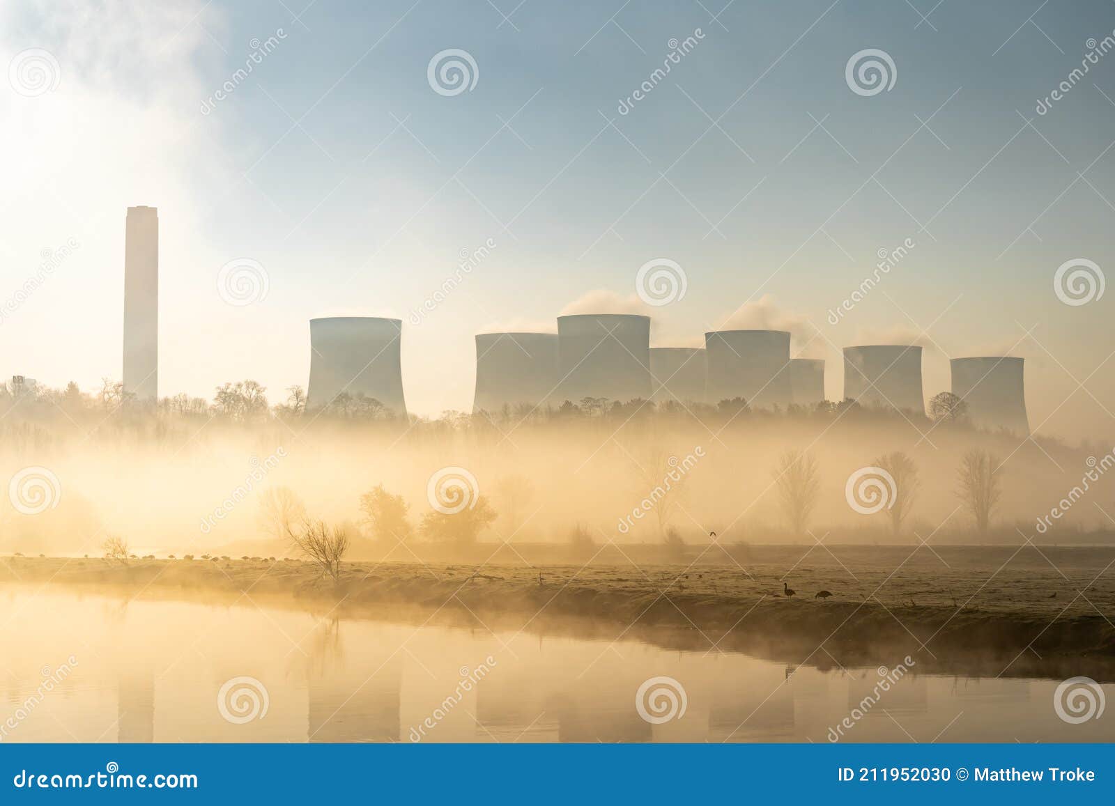 coal fired electric power station with chimneys and cooling towers surrounded by fog and mist at sunrise next to river haze