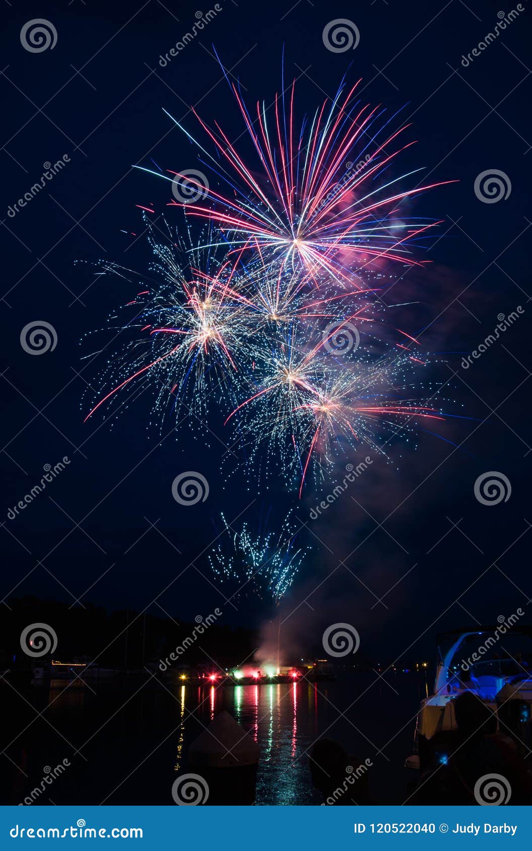 fireworks launched from a river barge with lights reflected in the water.