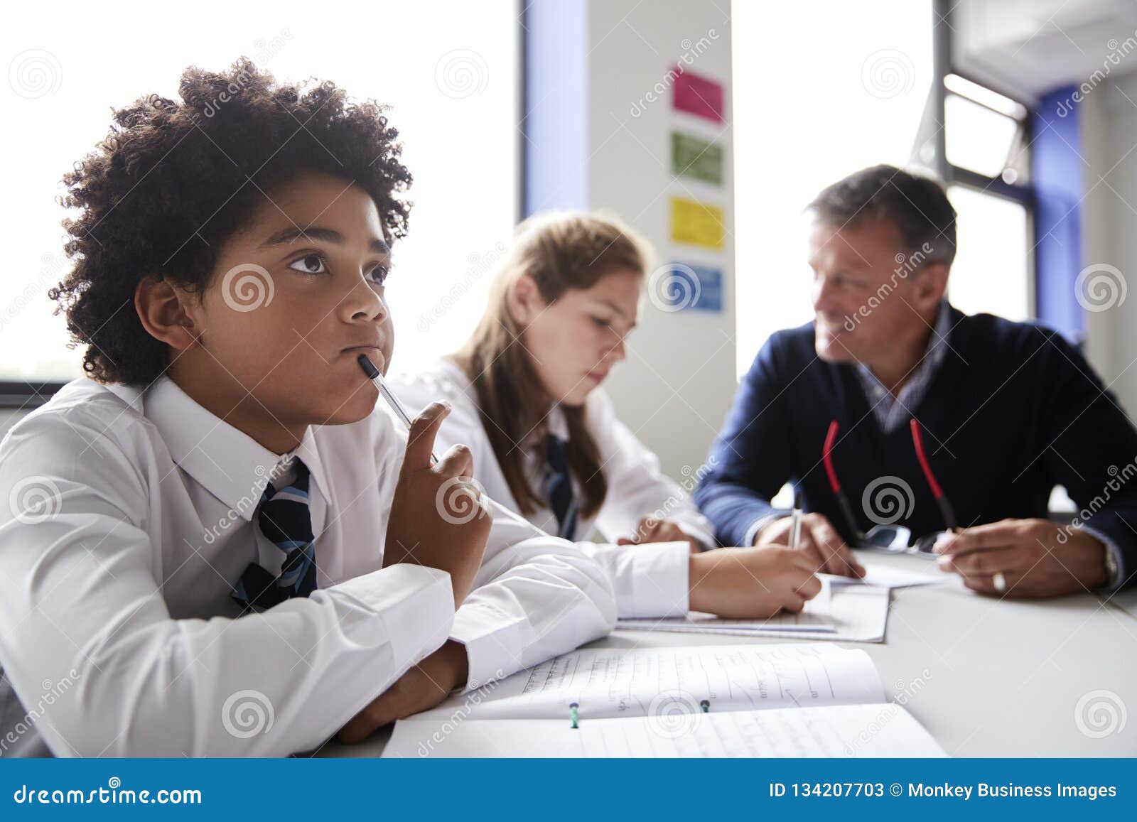 concentrating male high school student wearing uniform working at table with teacher talking to pupils in background