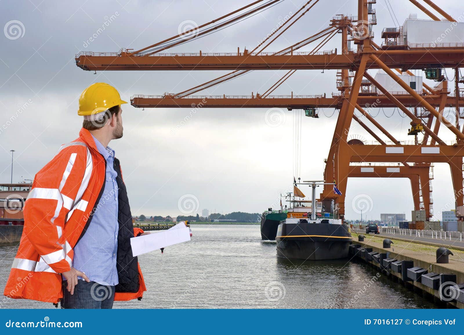 Controllo del porto. Un uomo che portano un cappotto di sicurezza e un cappello duro che controlla le gru enormi ad un contenitore harbor