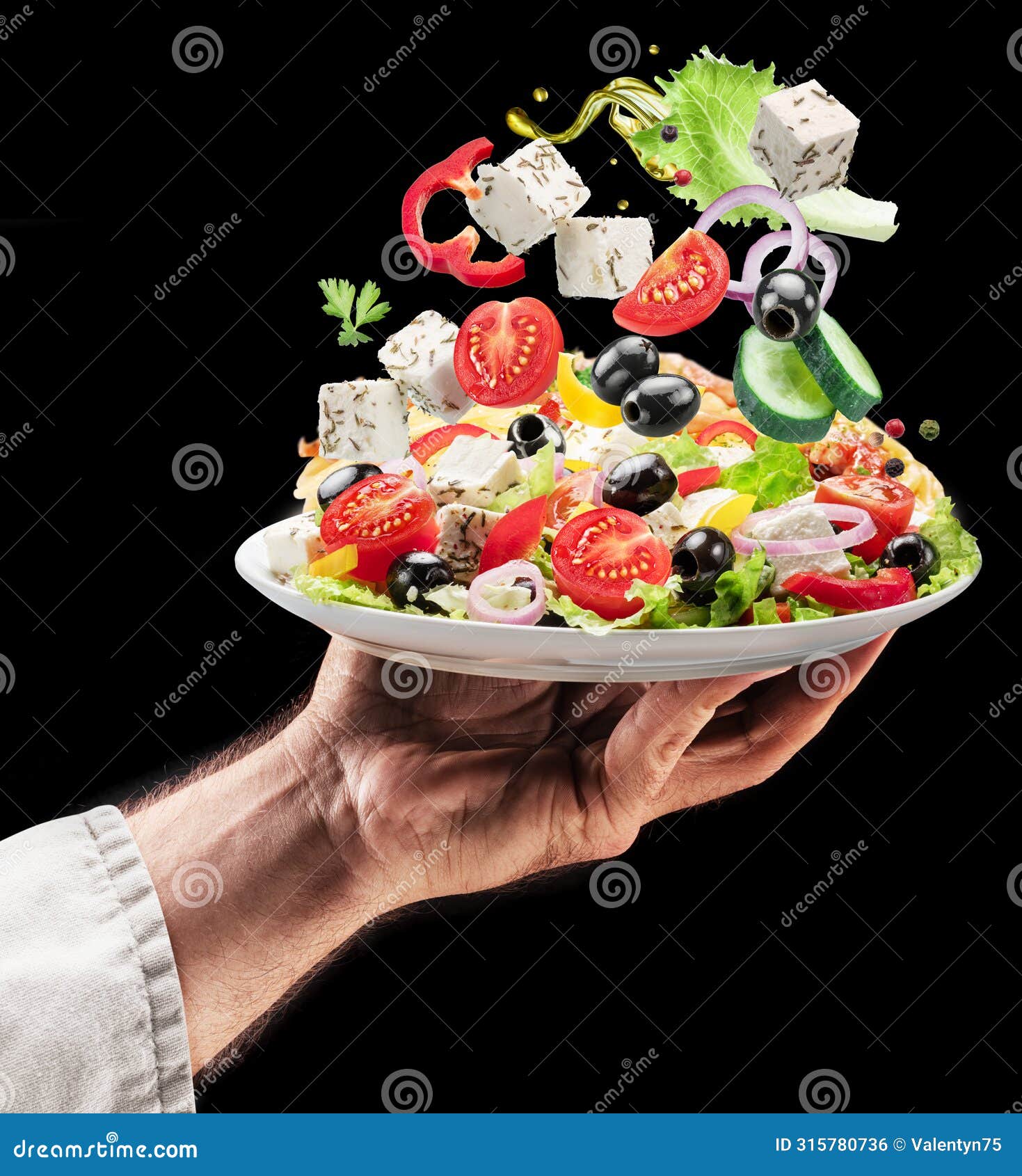 cook holds white plate of greek salad on black background. ingredients of salad falling down om the plate. tasty food background