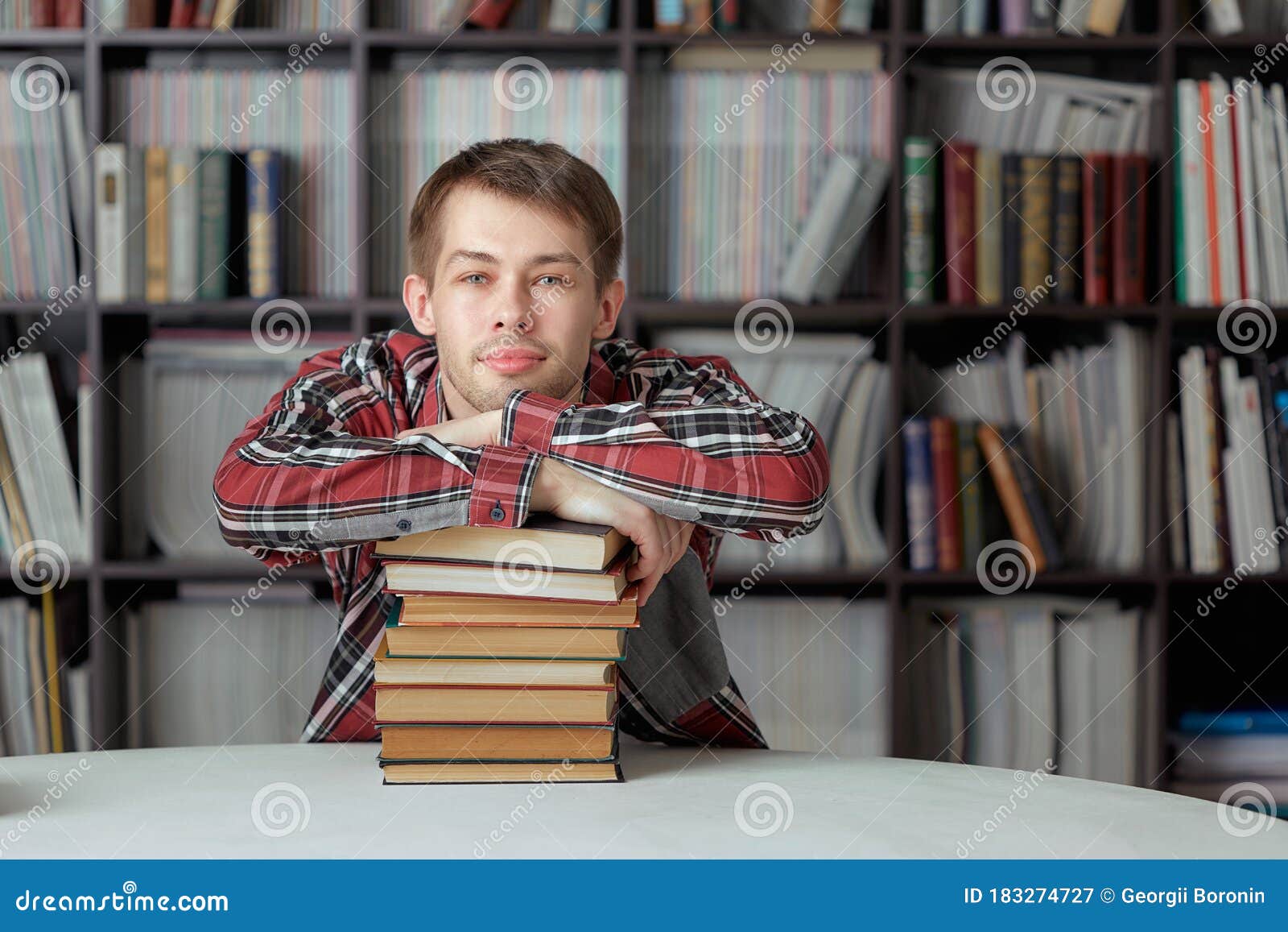 Cool Guystudent with a Smile Sitting at the Library Table and Hugging a Stack of Books