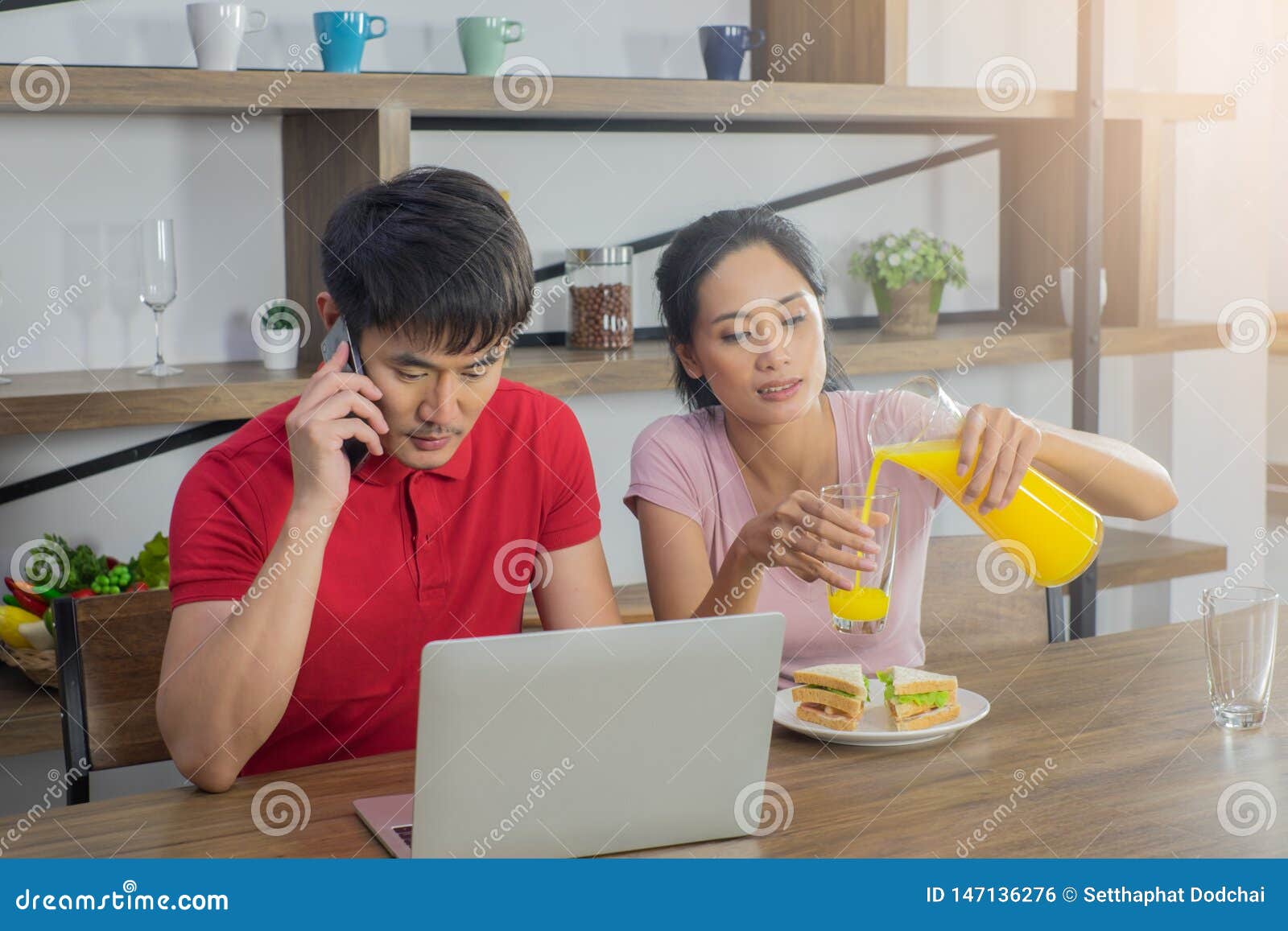 Coppie asiatiche, sedentesi al tavolo da pranzo Gli uomini stanno esaminando il computer portatile e stanno parlando sul telefono Donne che versano succo d'arancia in un vetro Ed avendo un piatto di panino per la prima colazione sulla tavola