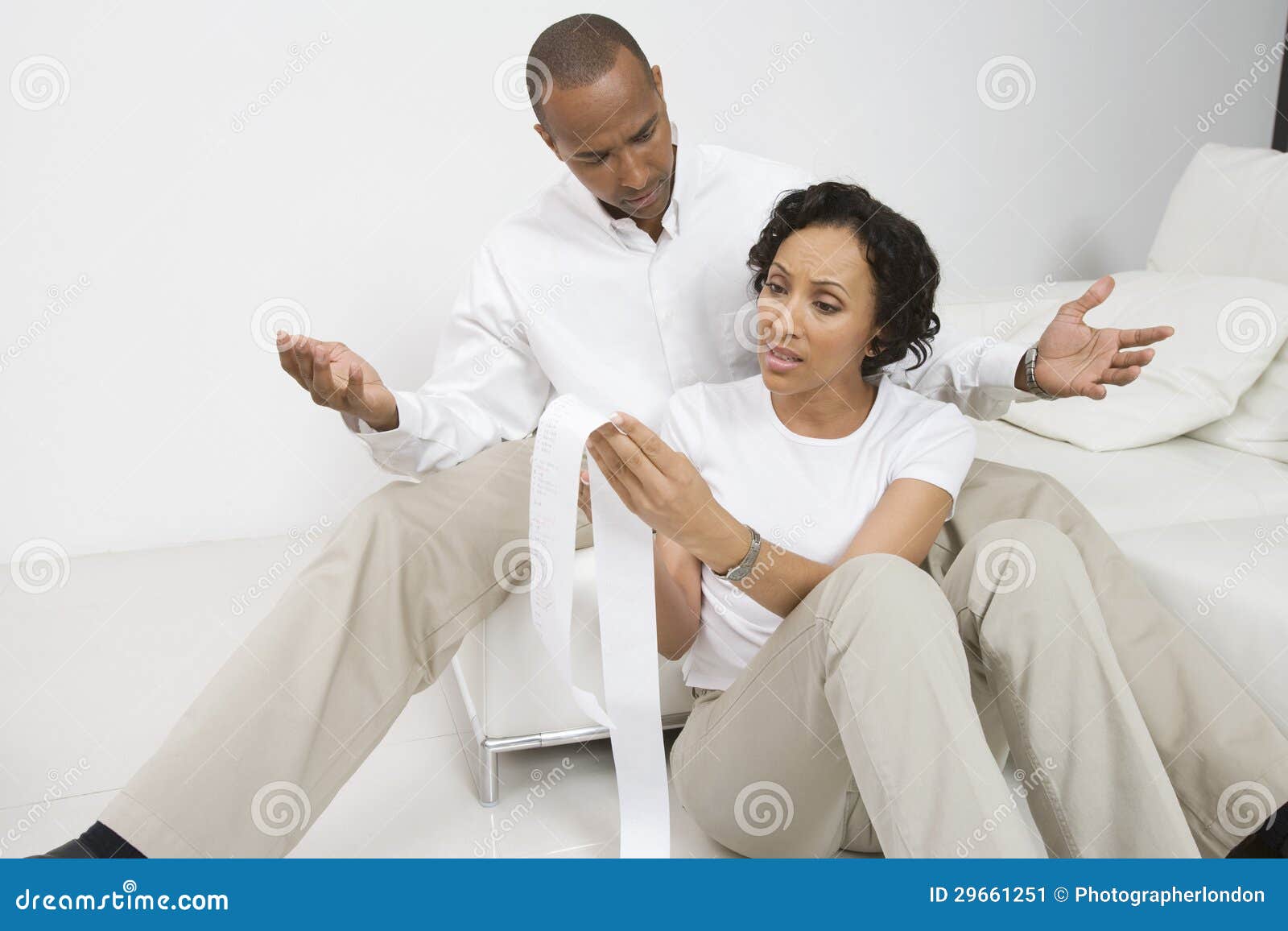  A man and a woman sit on the floor in front a couch looking at a long receipt with stressed expressions on their faces.