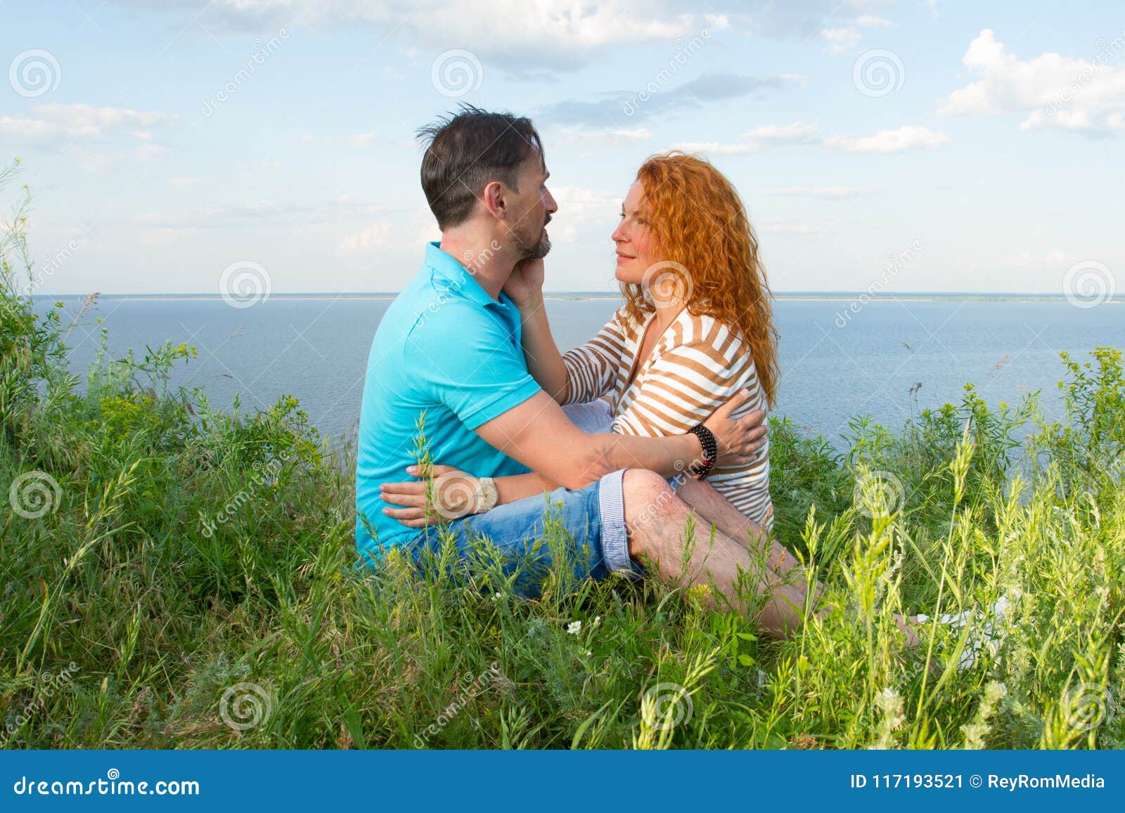 A Couple In Love Outdoor Lovers Sitting Hugged On The Grass At Lake Bank In Grass On Water And