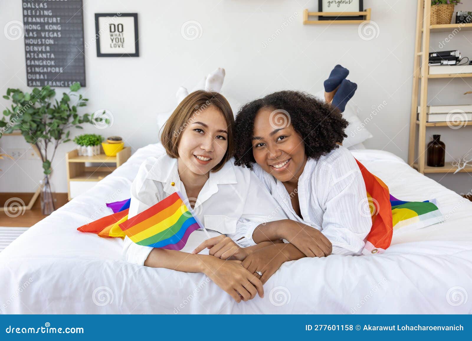 Couple of Same Sex Marriage from Difference Races Holding LGBTQ Rainbow Flag for Pride Month while Lying Down Together on the Bed Stock Photo image