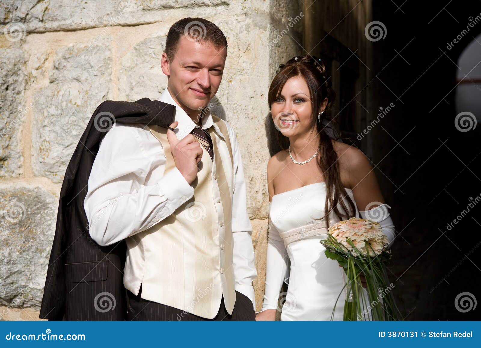 Couples nuptiales occasionnels. Mariée et marié se penchant sur un mur. Il utilise sa jupe occasionnelle au-dessus de son épaule.