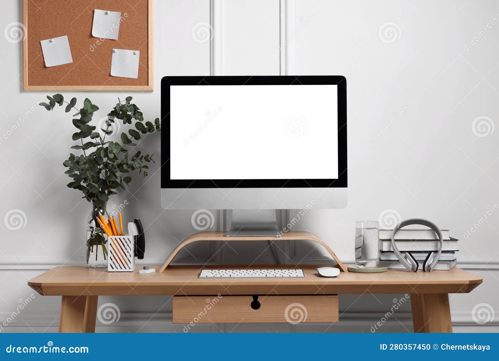 A desk with a computer, headphones, books, a glass of water, a vase with plants, a corkboard, and a pencil holder.
