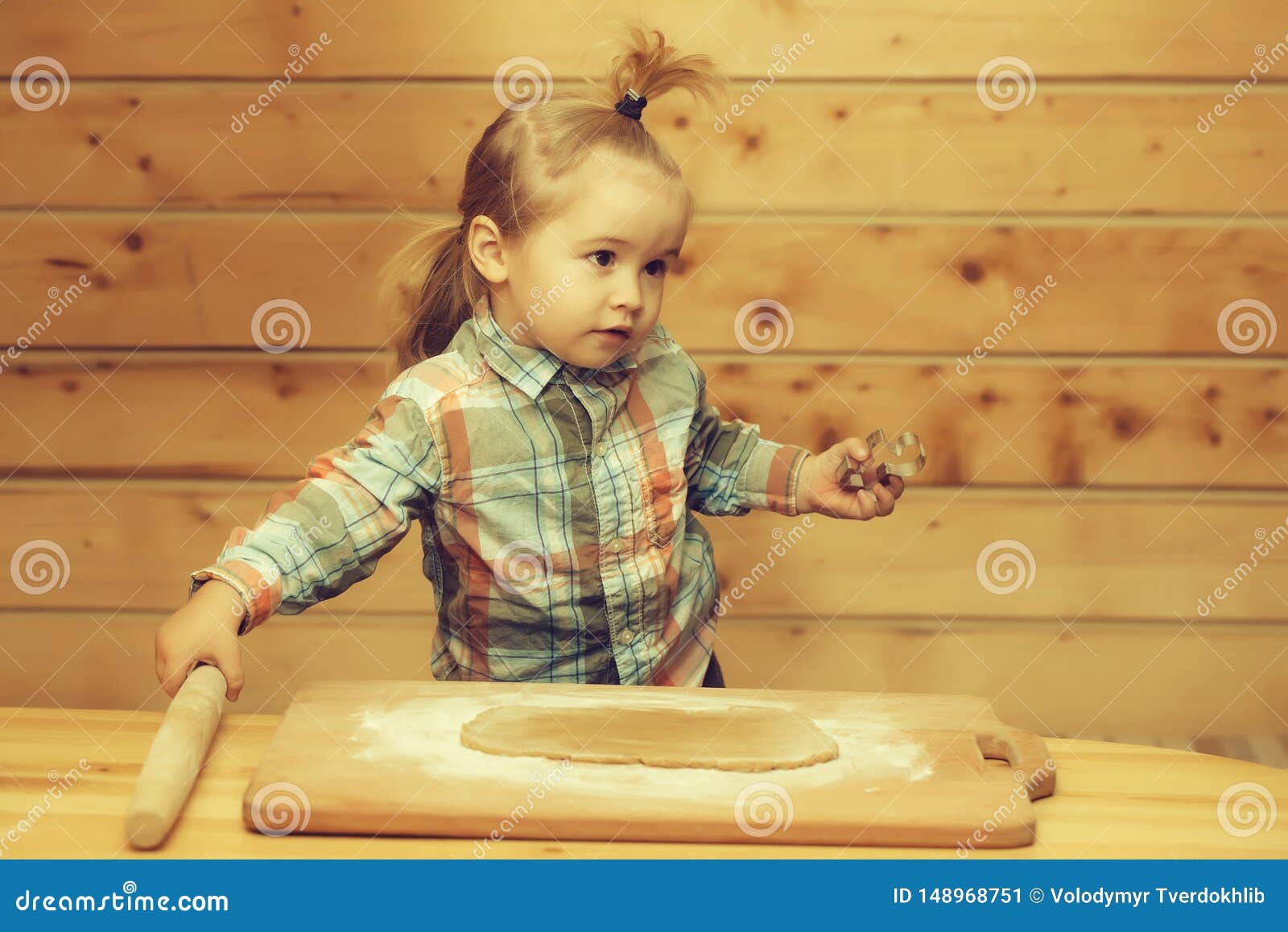 Crian?a bonito que cozinha com massa e farinha, molde met?lico das posses. Adorable small child chef or cute baby boy in fashionable chekered shirt cooking on board flour and dough holds metallic mold for cookies on wooden background.
