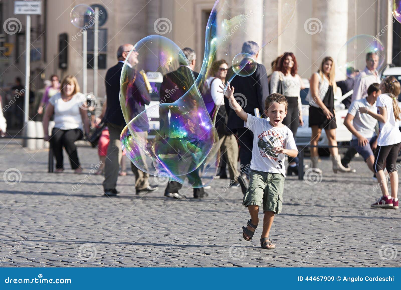 Criança feliz que corre para uma bolha de sabão. Uma criança feliz está correndo para alcançar uma grande bolha de sabão Atrás dele há muitos turistas em Praça del Popolo em Roma (Itália)