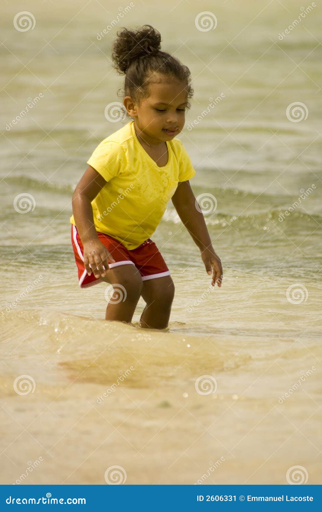 Criança pequena que vadeia na água. Brincadeiras fêmeas pequenas na água perto da linha costeira. A criança está desgastando shorts e t-shirt do verão e é da ascendência africana.