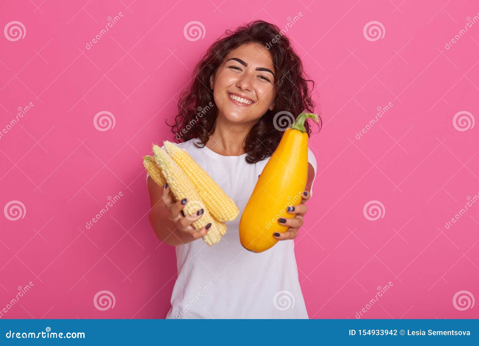 cropped image of young woman dressed in white casual t shirt showing zucchini and corncob to camera, has happy facial expresion,