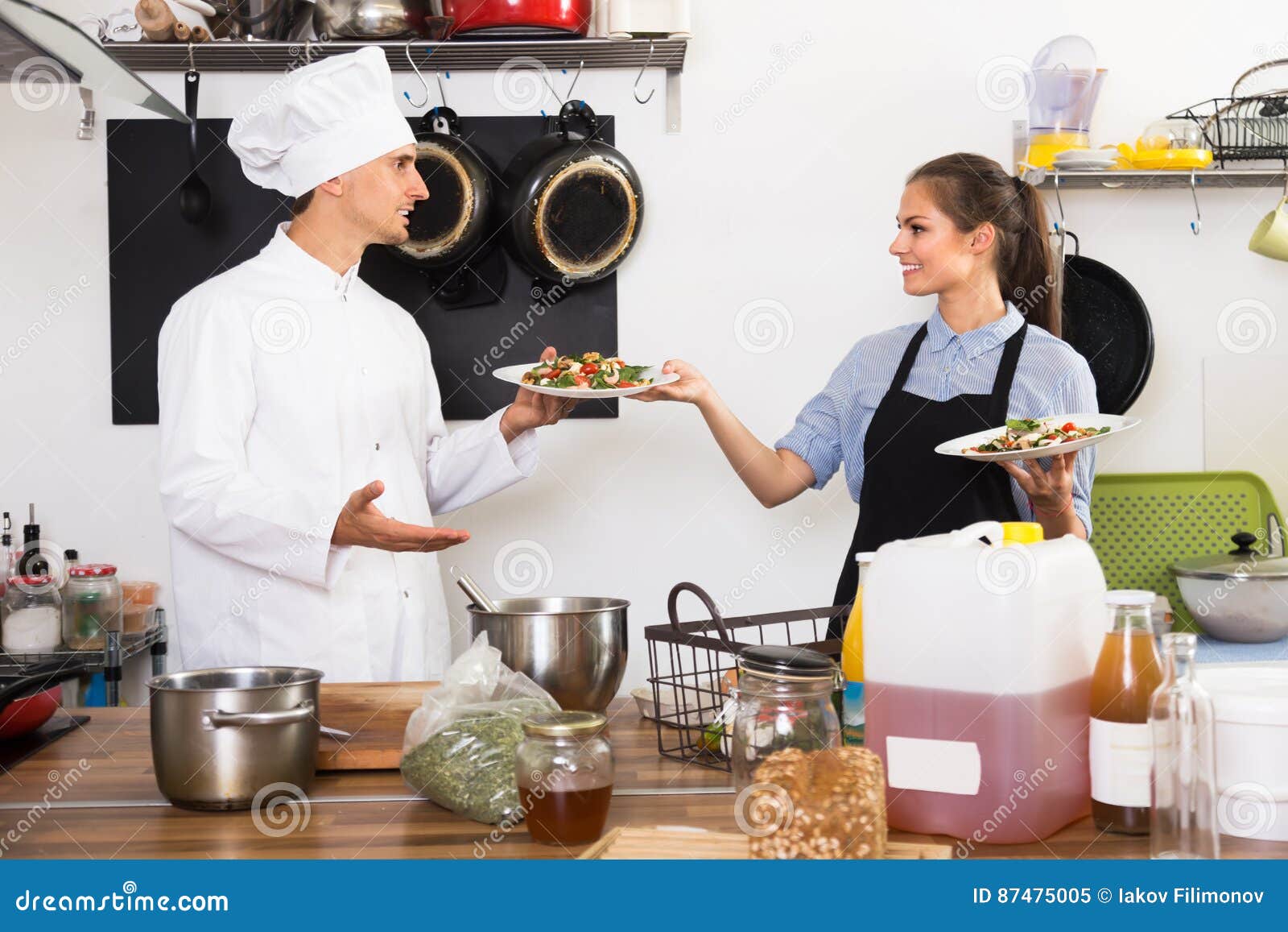 Cuoco allegro dell'uomo che dà insalata alla cameriera di bar. Gli uomini allegri cucinano nell'insalata del servizio dell'uniforme di bianco e nel darla alla cameriera di bar alla cucina del ` s del ristorante