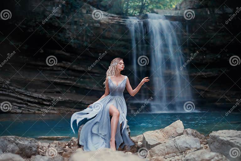 A Curly Blonde Girl in a Luxurious Blue Dress Sits on White Stones ...