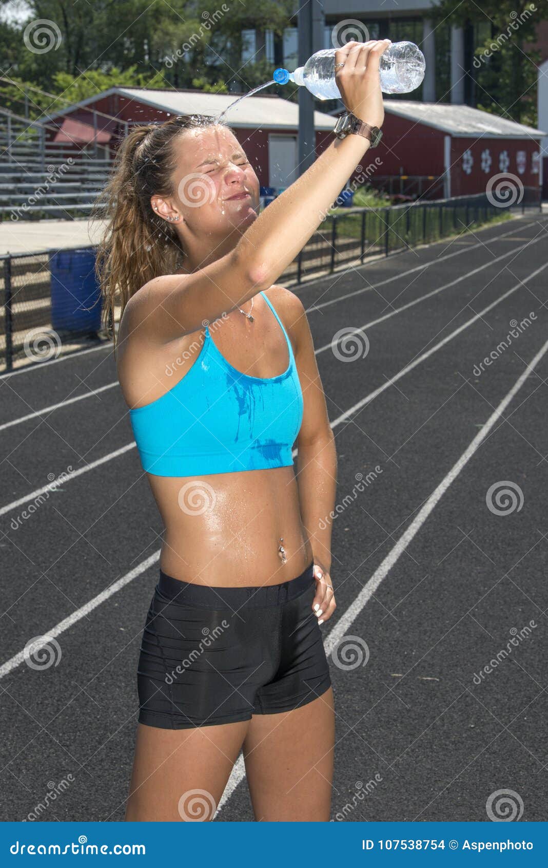 Cute and fit young teen Caucasian girl working out on outdoor track  glistens with sweat in blue sports bra as she pours water on her face Stock  Photo