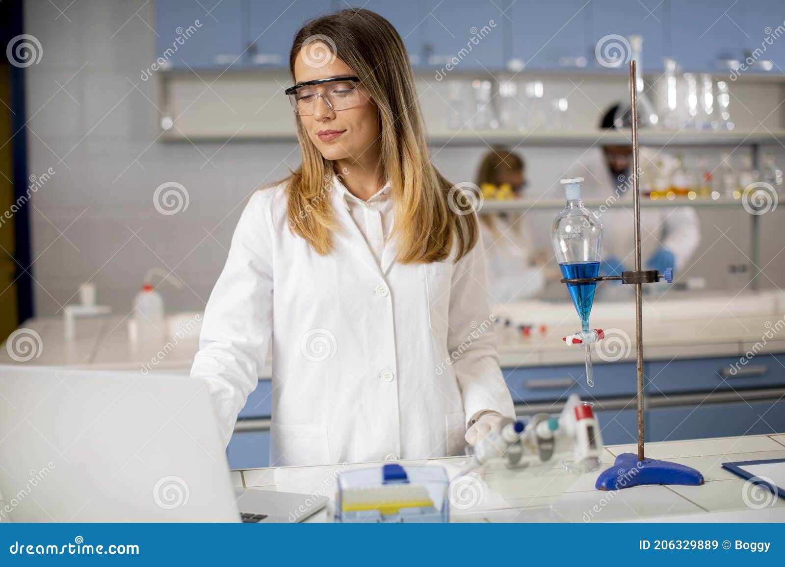 Female Researcher in White Lab Coat and Protective Mask Using Laptop