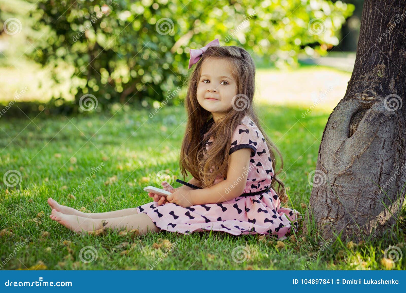 cute girl sitting on green grass close to old tree barefoot during summer time vacation in awesome stylish pink dress with cell ph