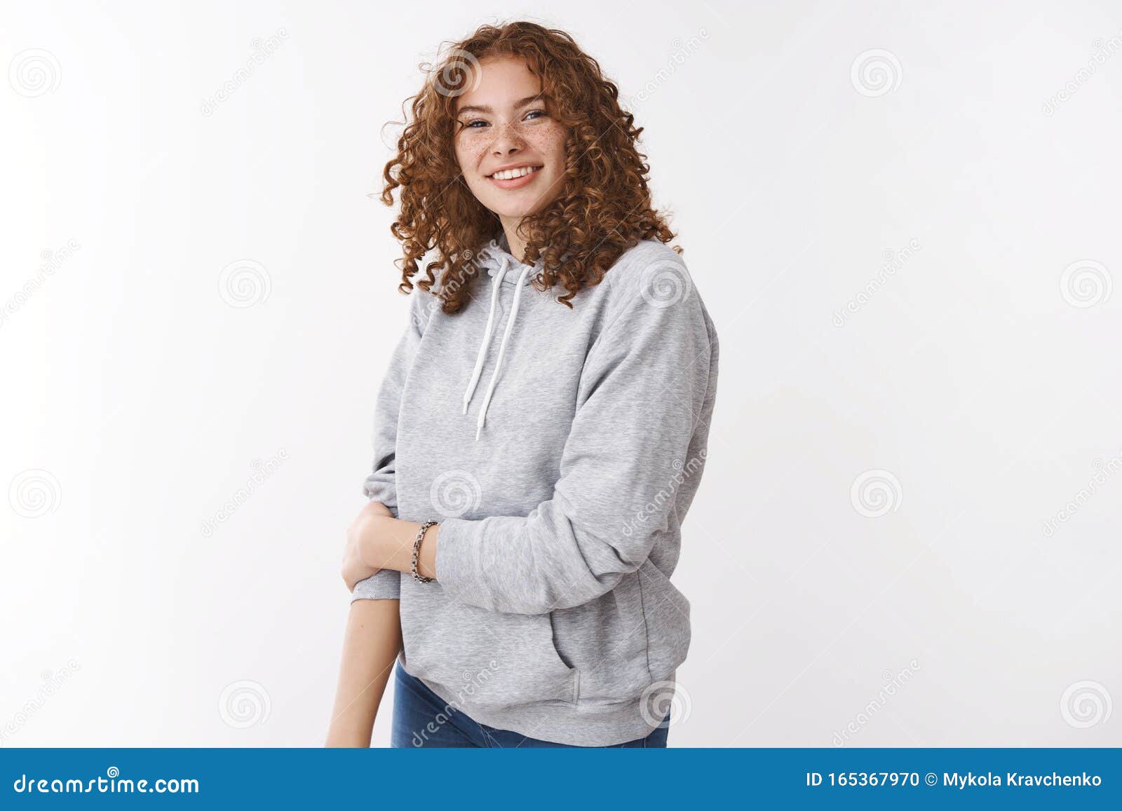 cute insecure young redhead teenage girl with freckles curly hair wearing grey hoodie smiling touch arm unconfident
