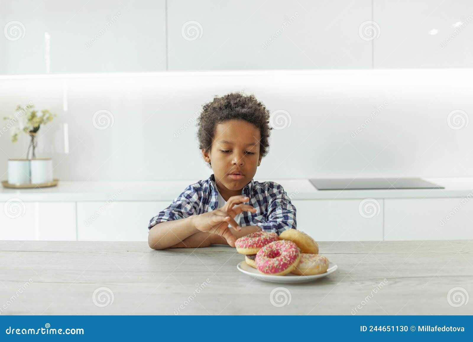 Cute Kid Boy with Black Hair Eating Donuts in White Kitchen Stock Photo