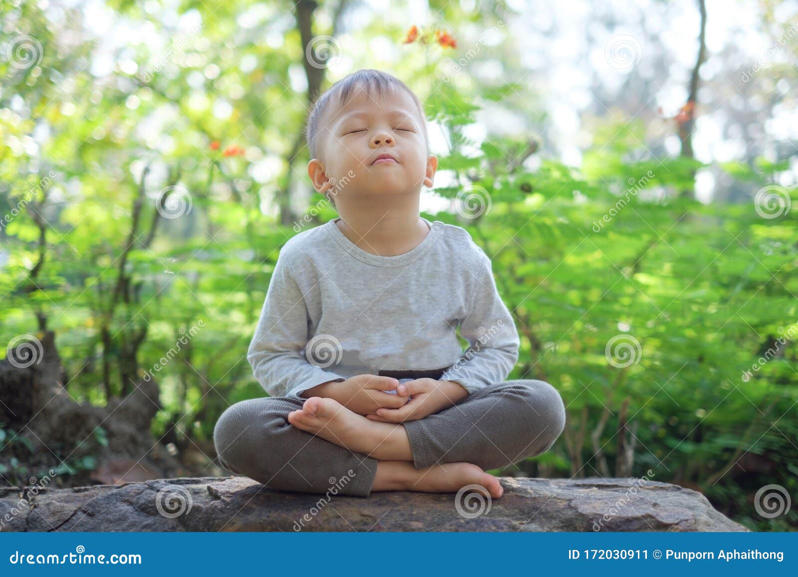 cute little asian 2 - 3 years old toddler baby boy child with eyes closed, barefoot practices yoga & meditating outdoors on nature