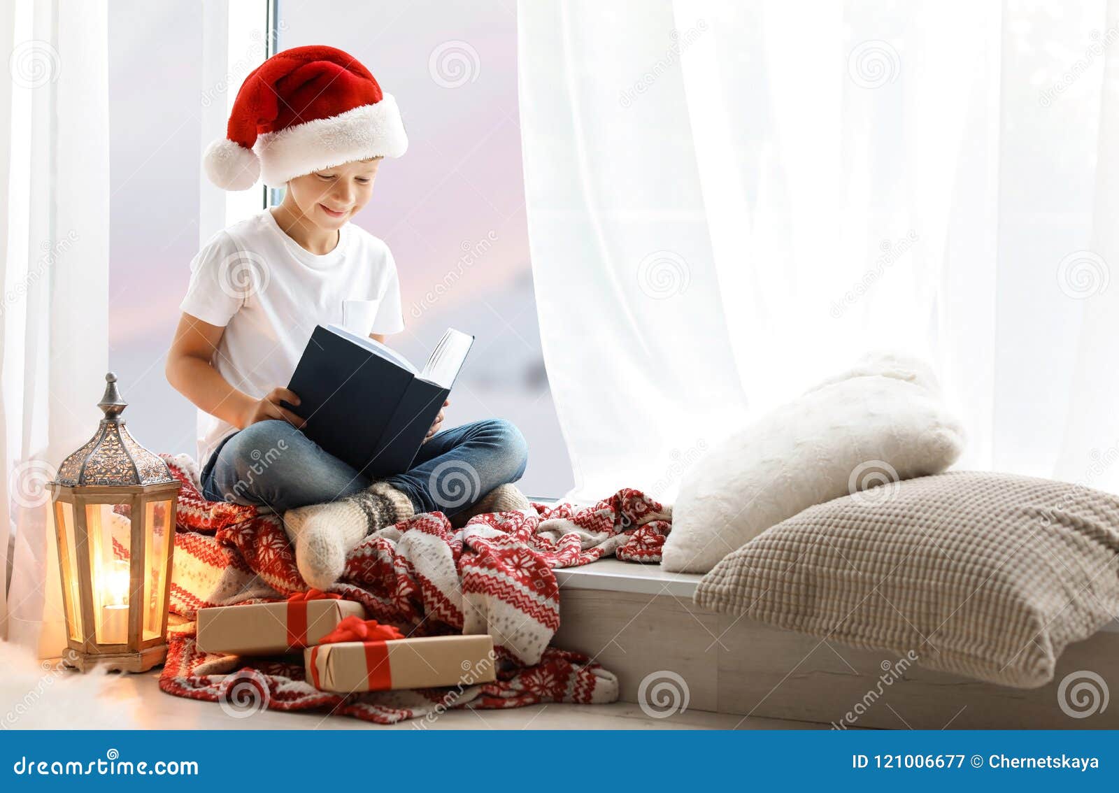 Cute little child in Santa hat reading Christmas story while sitting on windowsill