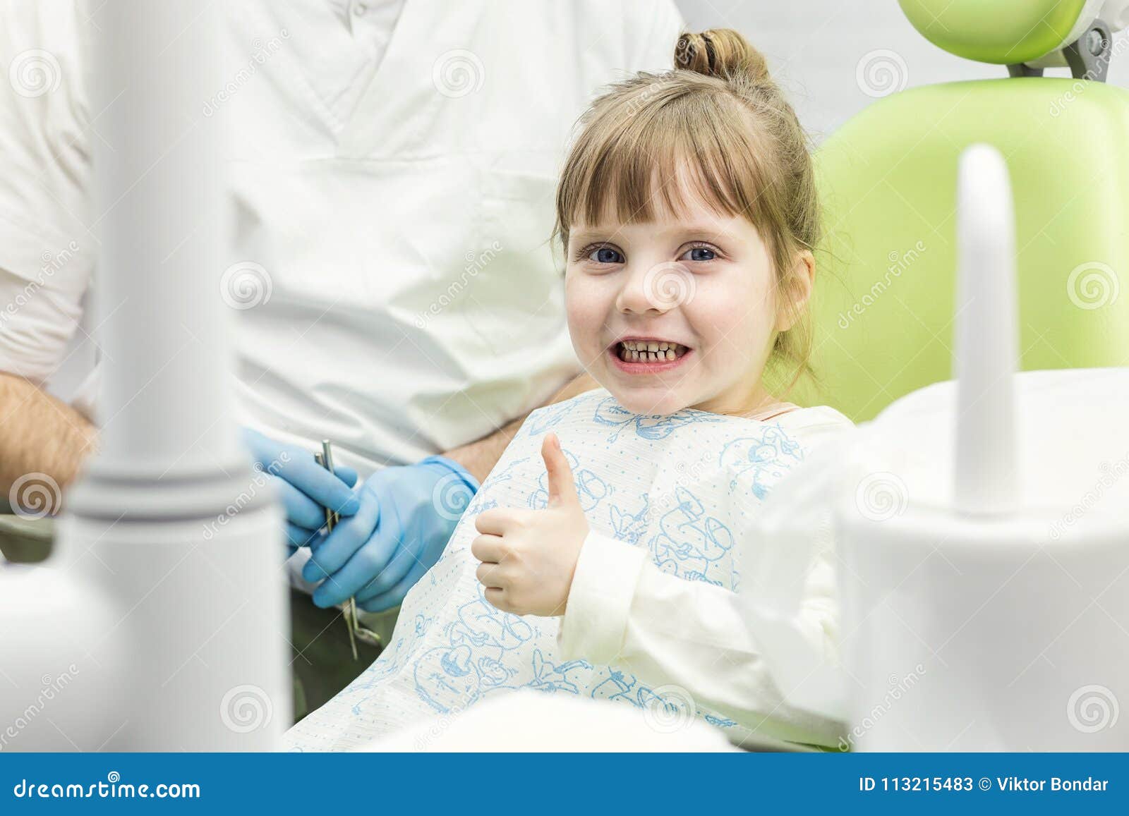 cute little girl showing thumb up sign at dentist`s office clin
