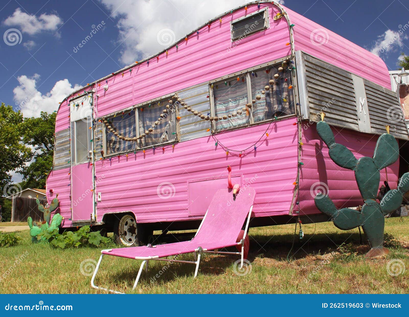 cute pink travel trailer under a bright cloudy sky with cacti and trees around a tumbona chair