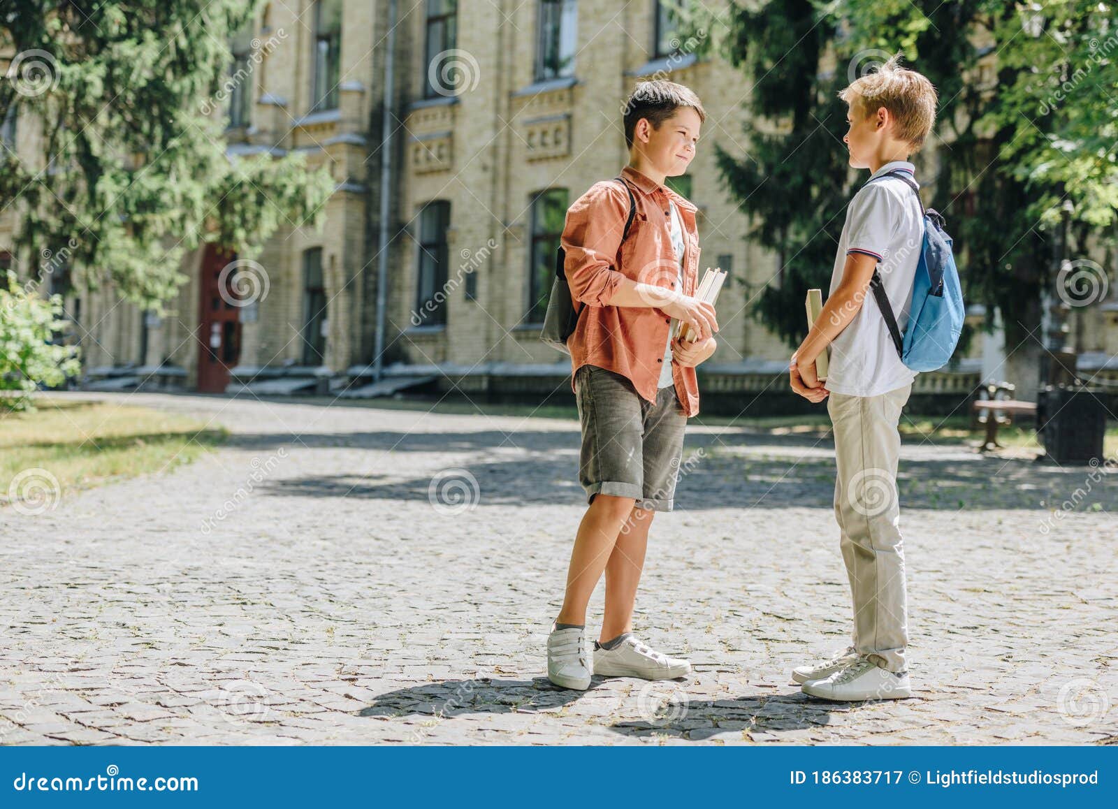 cute schoolboys holding books and speaking while standing in schoolyard