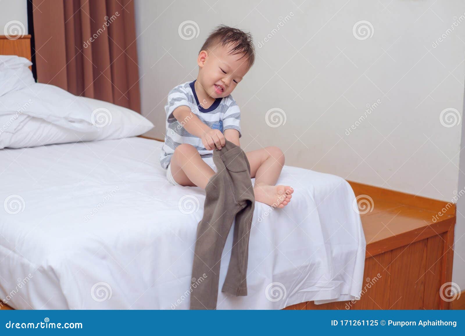 cute smiling little asian 2 years old toddler boy child sitting in bed concentrate on putting on his pants