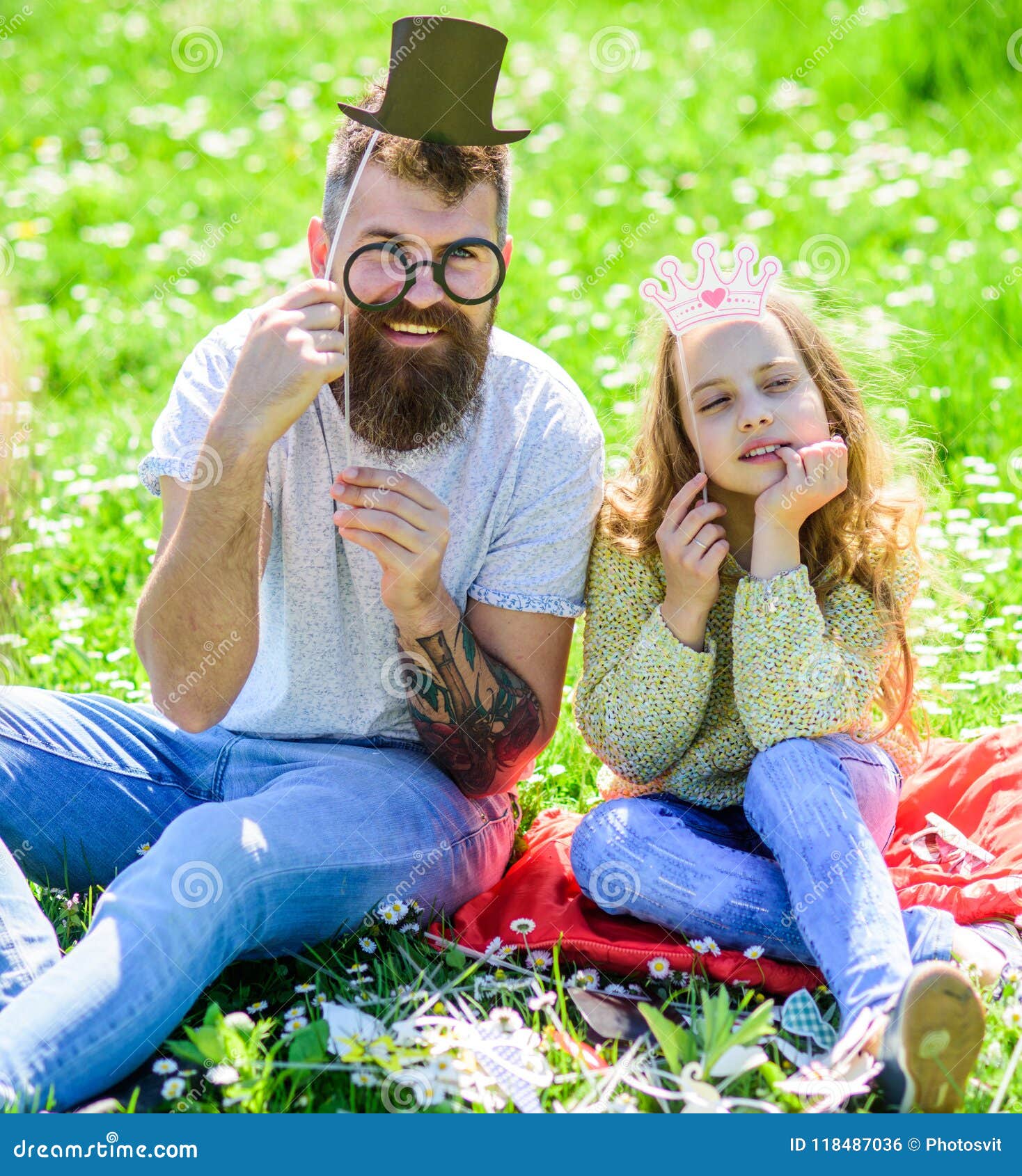 dad and daughter sits on grass at grassplot, green background. child and father posing with eyeglases, crown and top hat
