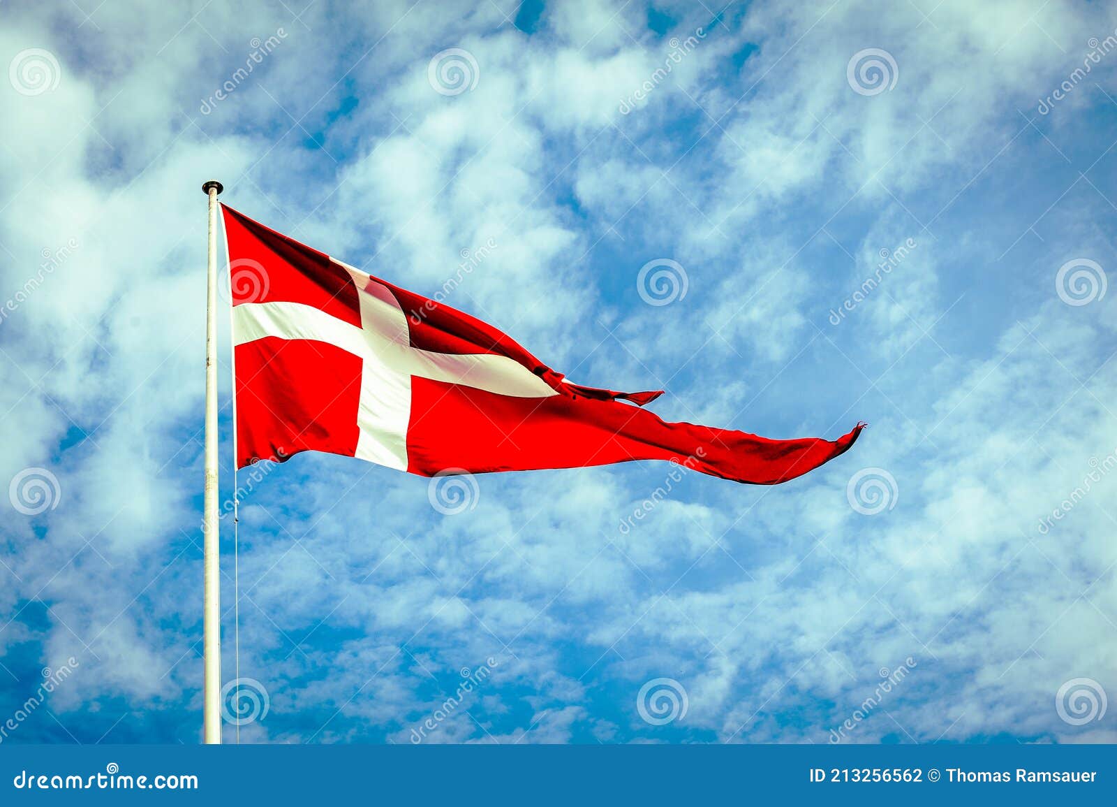 danish flag, the dannebrog, in front of a blue and cloudy sky.