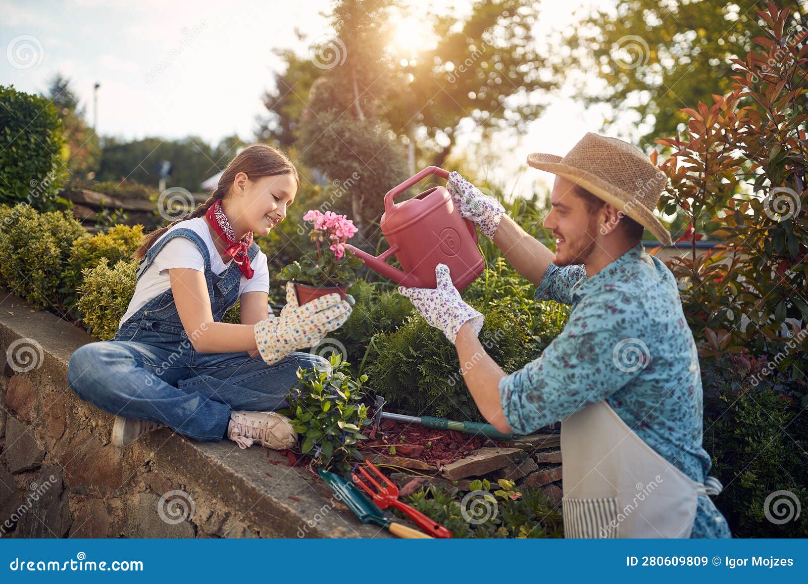 daugther and father working together in the flower garden outdoors, girl holding a flower pot, man watering it with watercan